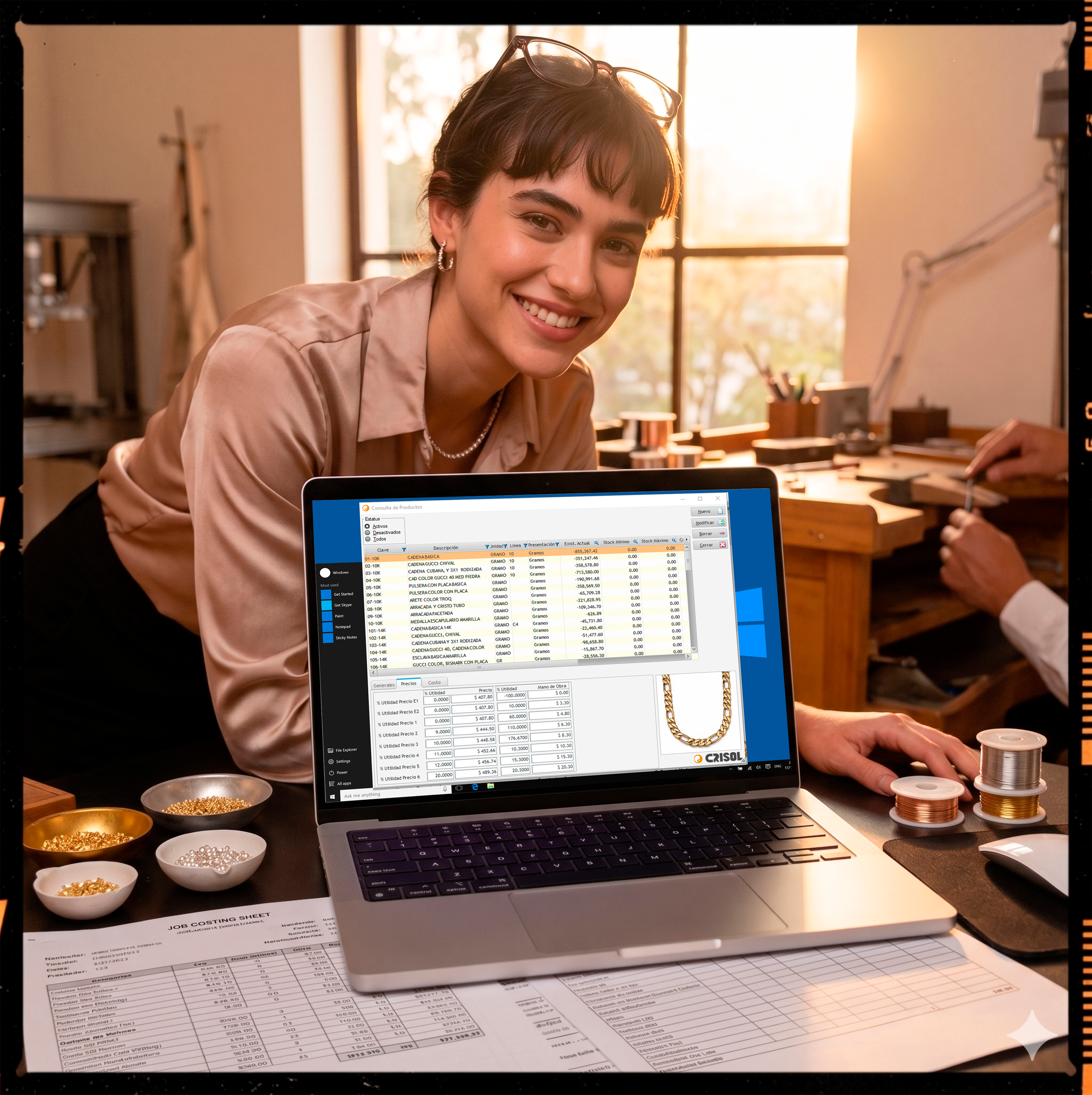 Laptop en un taller de joyería mostrando una pantalla de traspasos de mercancía y existencias por sucursal, rodeada de rollos de metal y una orden de costo, representando la logística interna y trazabilidad.
